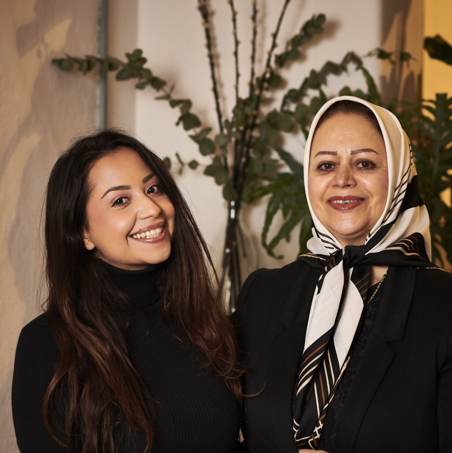 Two women smiling, standing side by side indoors. The younger woman wears a black top, and the older woman wears a black outfit with a patterned headscarf. A plant is visible in the background.